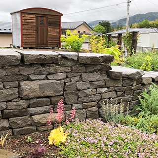 庭の花壇を支える手積みの石積み土留め。濃い灰色の自然石を積み上げ、物置や遠景の山並みが見える造園の一例。